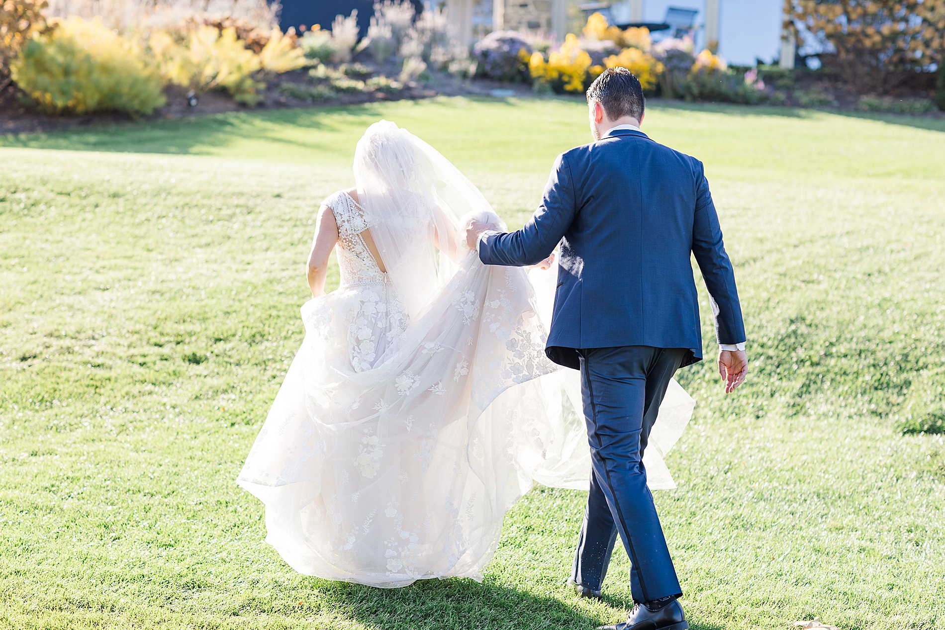 groom carries train of bride's dress as they walk across the lawn