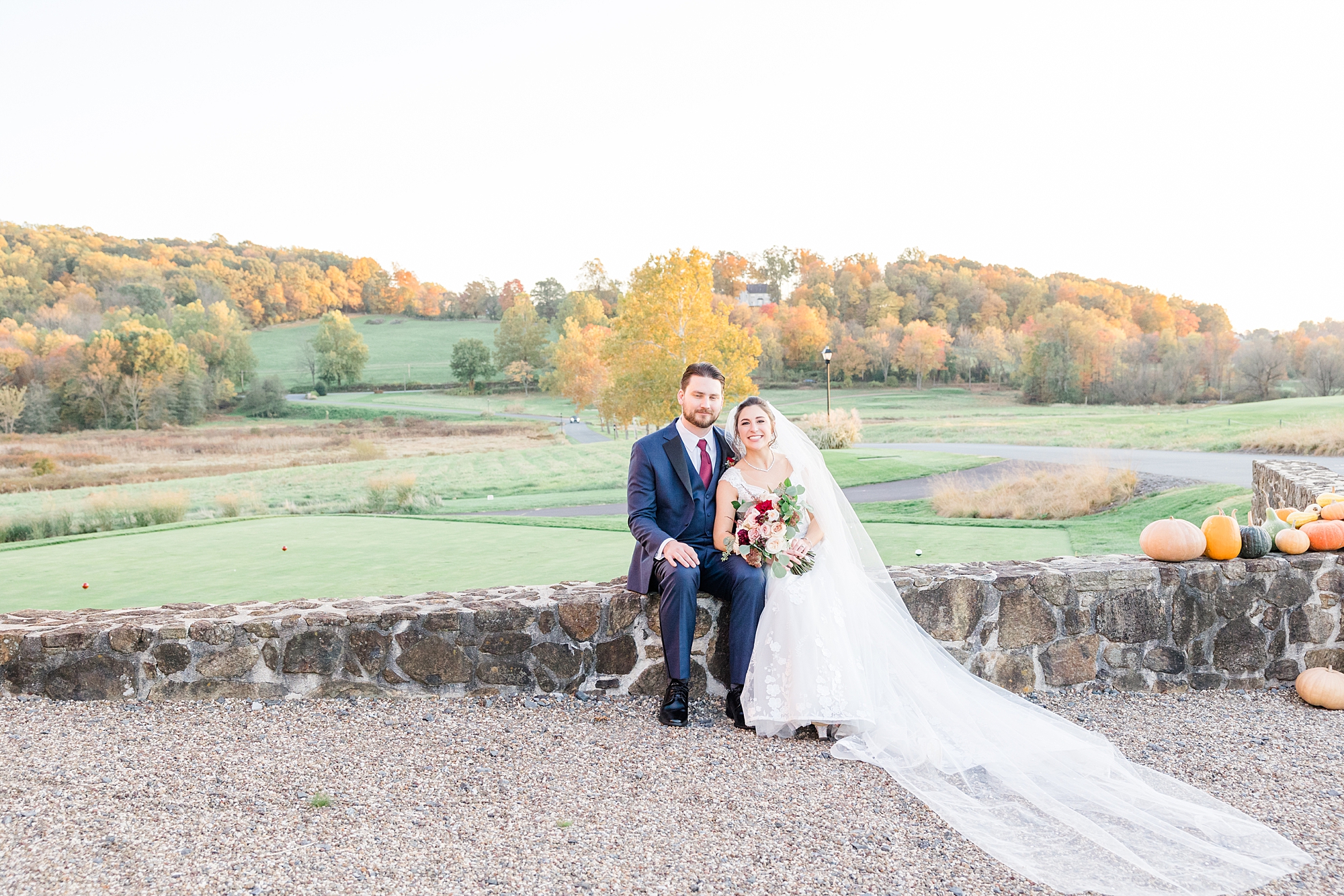 bride and groom sit on stone wall with the fall trees in the background