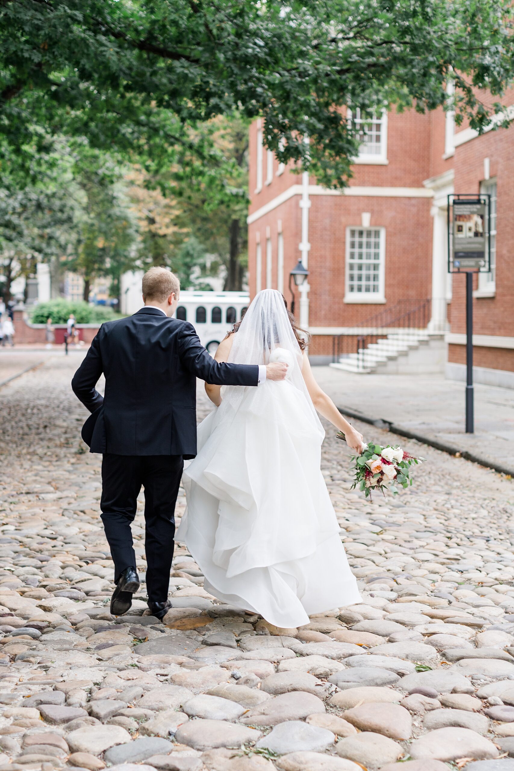 groom holds up bride's dress as they walk down cobblestone street